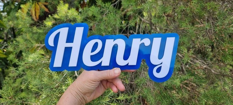 Op de afbeelding: Een blauw acryl bord met de naam "Henry" geschreven in witte cursieve letters.