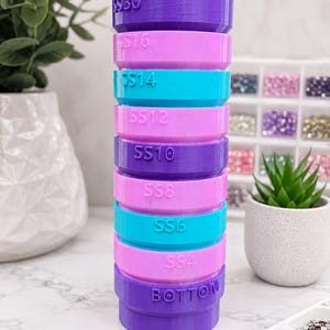 May include: A vertical stack of cylindrical containers in alternating shades of blue, purple, and pink. Each container is labeled with text including TOP, SS30, SS16, SS14, SS12, SS10, SS8, SS6, SS4, and BOTTOM, indicating sizes for sorting small craft items.