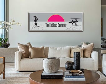 Puede incluir: Un cuadro enmarcado que presenta una escena de playa con dos figuras silueteadas que llevan tablas de surf. Un sol rosa se pone sobre el océano, con las palabras "The Endless Summer" debajo. La obra de arte se exhibe sobre un sofá blanco.