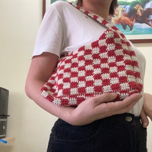 May include: A red and white checkered crochet bag with a long strap. The bag is being held by a person.
