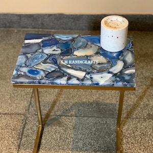 May include: A gold metal side table with a blue agate stone top. The table has a square base and a rectangular top. The agate stone is a natural material with a variety of blue and white colours. The table is ideal for a living room or bedroom.