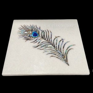 Puede incluir: Posavasos cuadrado de mármol blanco con un diseño de pluma de pavo real incrustado con nácar iridiscente y piedras azules y turquesas. El posavasos tiene el texto "N.M. HANDICRAFTS" en la parte inferior.