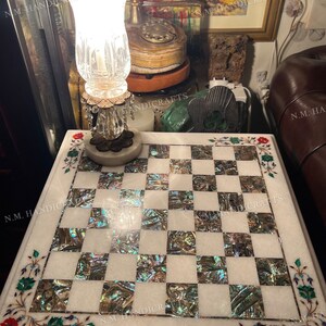 May include: A white marble chessboard inlaid with mother of pearl and floral designs. The chessboard is on a table with a crystal lamp and a small wooden box.