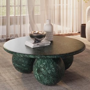 Puede incluir: Una mesa de centro de mármol verde con tapa redonda y tres patas. La mesa está sobre una alfombra marrón claro en una sala de estar con un sofá blanco y una planta en maceta.
