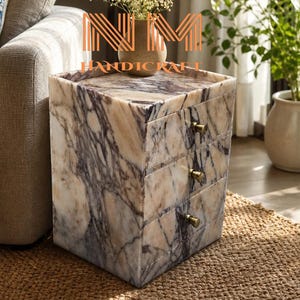Pode incluir: Uma mesa de cabeceira quadrada com um padrão de mármore branco e roxo, contendo três gavetas, cada uma com um pequeno puxador cor de latão. O texto NM HANDICRAFT é exibido em letras laranja na superfície da mesa.