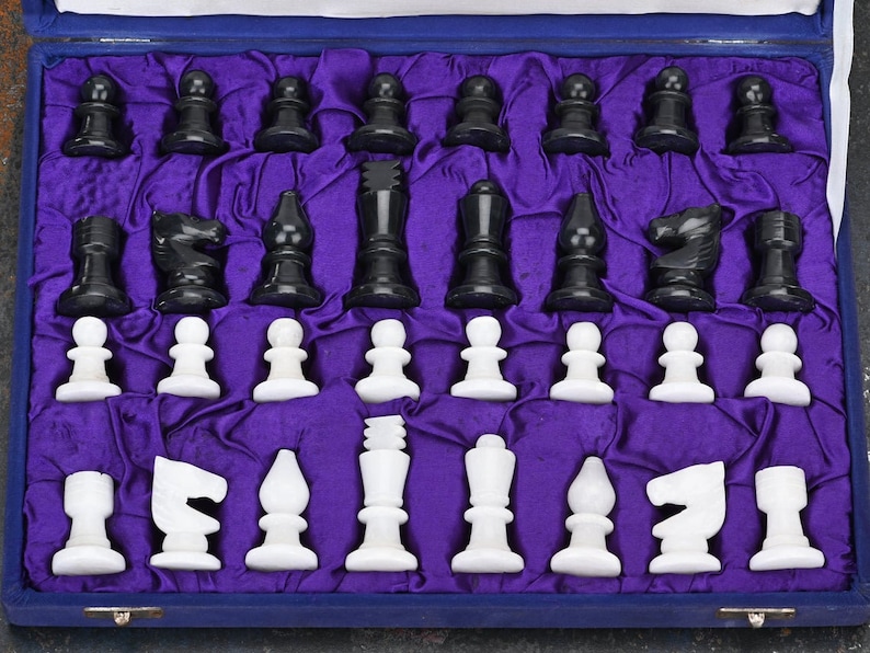 May include: A set of black and white chess pieces in a purple velvet lined box. The pieces are made of polished stone and are arranged in their starting positions.