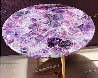 May include: A round table with a purple amethyst top and a gold metal base. The table top is made of many small pieces of amethyst that have been polished and glued together. The base is a simple, modern design with three legs.
