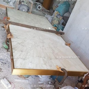 May include: Two white rectangular panels with a gold trim are being assembled. The panels are made of a white material with a textured surface. The panels are being held together by a clamp. The panels are being worked on in a workshop setting.