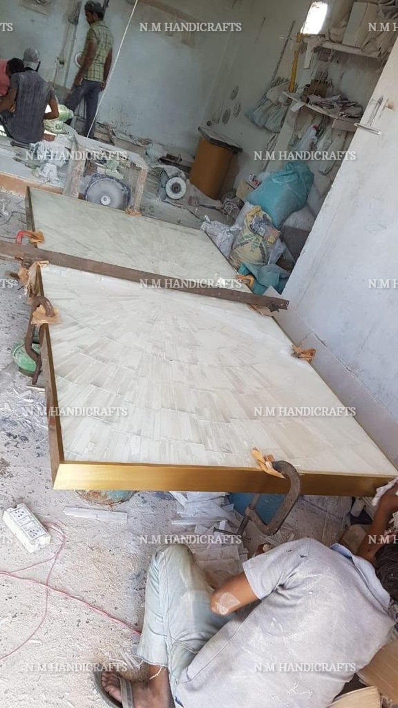 May include: Two white rectangular panels with a gold trim are being assembled. The panels are made of a white material with a textured surface. The panels are being held together by a clamp. The panels are being worked on in a workshop setting.