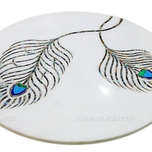 Puede incluir: Plato redondo de mármol blanco con dos plumas de pavo real incrustadas con nácar y detalles azules y verdes. El plato tiene un diámetro de 30 cm.