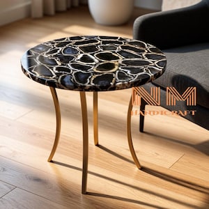 May include: A round side table featuring a tabletop composed of polished black and white agate slices with a smooth finish. The table is supported by three slender, curved gold-toned metal legs. The text NM HANDICRAFT is visible in orange.