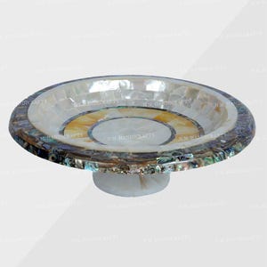 May include: A decorative, round, pedestal bowl. The bowl features a mosaic design with white, yellow, and iridescent shell pieces. The base is white. The bowl is likely used for display or serving.