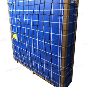 May include: A large rectangular cardboard shipping box wrapped in a black cargo net. The box features a bright blue surface with a white grid pattern, brown edges, a small yellow square label, and the text N.M. HANDICRAFTS printed repeatedly.