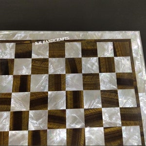 May include: A chessboard made of tiger eye and mother of pearl. The board is a standard 8x8 grid with alternating squares of white mother of pearl and brown tiger eye. The board is surrounded by a white mother of pearl border.