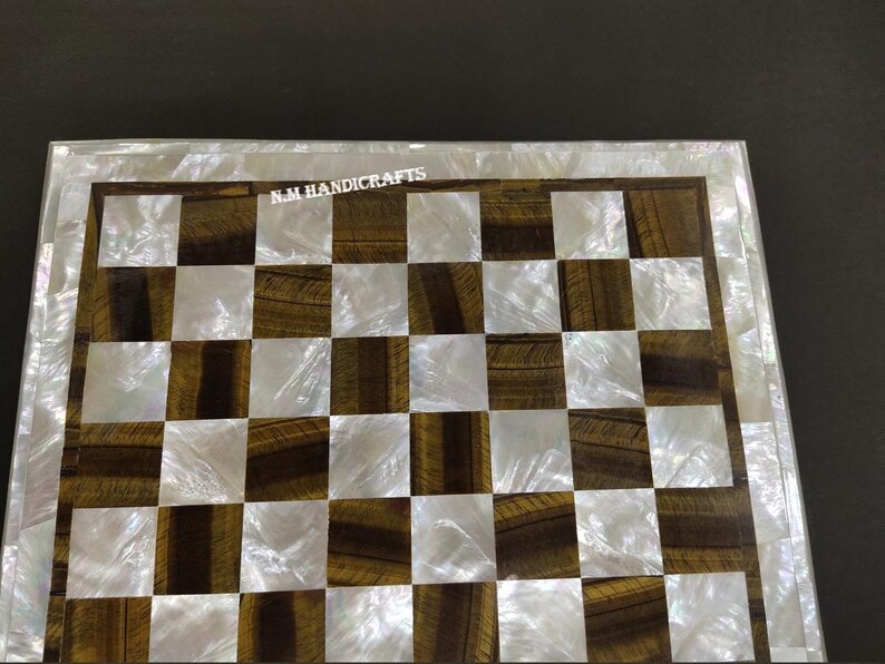 May include: A chessboard made of tiger eye and mother of pearl. The board is a standard 8x8 grid with alternating squares of white mother of pearl and brown tiger eye. The board is surrounded by a white mother of pearl border.