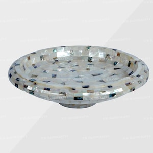 May include: A round serving tray with a mosaic design. The tray has a raised edge and a pedestal base. The mosaic is made of white and iridescent pieces, creating a shimmering effect. The tray is suitable for serving food or as a decorative item. The diameter is approximately 25 cm.