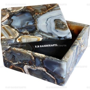 May include: A square agate box with a lid. The box is made of natural agate stone with a variety of colors, including gray, brown, and white. The lid is open and the inside of the box is black.