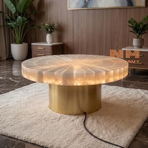 May include: A round coffee table featuring a translucent, white, sunburst-patterned stone top that emits a warm internal glow. The table is supported by a cylindrical, brushed gold metal base and includes a black power cord extending from the bottom.