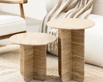 May include: Two round travertine side tables with a unique, geometric base. The tables are made of natural stone and have a warm, earthy tone.