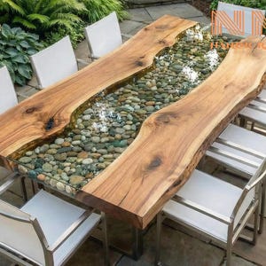 May include: A rectangular dining table featuring two natural live-edge wood slabs separated by a central glass-topped channel filled with smooth, multi-colored river stones, surrounded by six chairs with white fabric seats and metal frames.