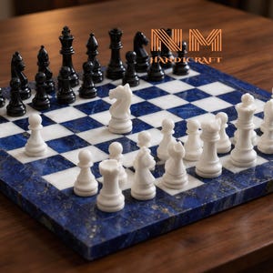 Könnte beinhalten: Ein quadratisches Schachspiel mit einem Brett aus abwechselnd dunkelblauen und weißen Steinfeldern. Das Set enthält einen vollständigen Satz geschnitzter schwarzer und weißer Schachfiguren, die spielbereit aufgestellt sind. Der Text NM HANDICRAFT ist in Gold zu sehen.