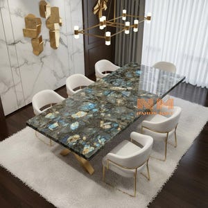 Peut inclure: Une table à manger rectangulaire avec un plateau en pierre labradorite polie bleue et grise sur une base en métal doré en forme de croix, entourée de six chaises blanches rembourrées à structure dorée. Le texte NM HANDICRAFT est visible.