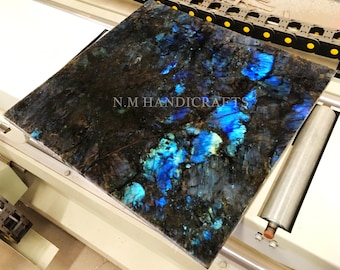 May include: A close-up of a large, polished slab of labradorite gemstone. The stone has a dark blue and black background with iridescent blue and green flashes. The text "N.M HANDICRAFTS" is visible in the lower left corner of the image.