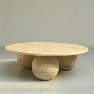 May include: A round, light beige travertine coffee table with three spherical legs.