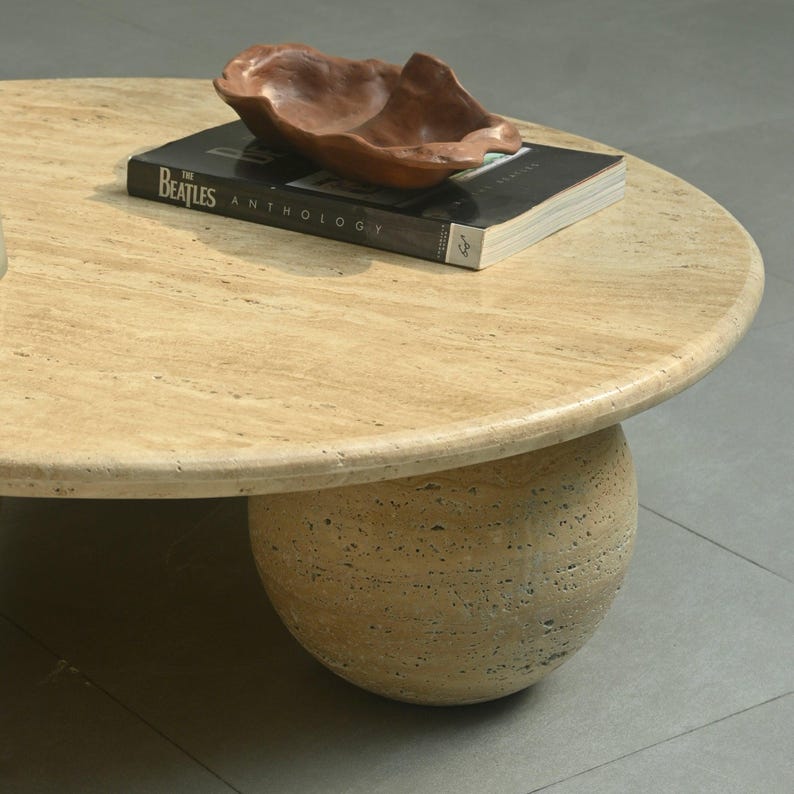 May include: A round travertine coffee table with a smooth, light brown top and three rounded legs. A book titled "The Beatles Anthology" and a wooden bowl are on top of the table.