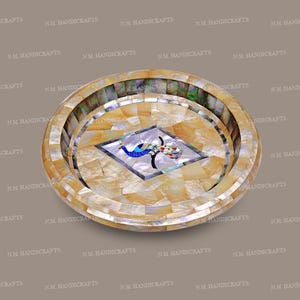May include: A decorative, round tray with a mosaic design. The tray features a central peacock motif in blue, red, and green, surrounded by a geometric pattern of beige and white tiles. The outer rim has a layered design.