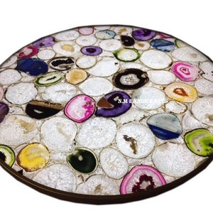 May include: Round table top inlaid with polished agate slices in various colors, including white, pink, purple, yellow, and green. The agate pieces are set in a white matrix, creating a mosaic effect. The table top has a dark brown border and the text "N.M HANDICRAFTS".