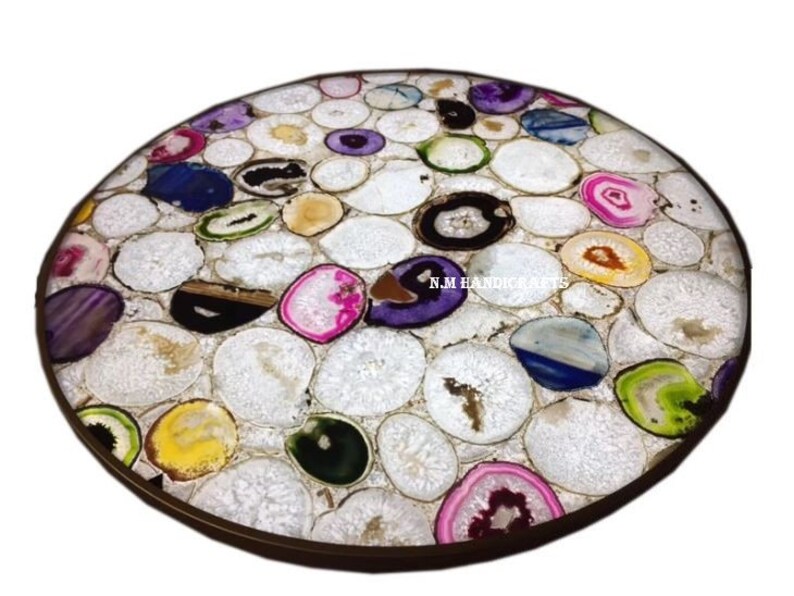 May include: Round table top inlaid with polished agate slices in various colors, including white, pink, purple, yellow, and green. The agate pieces are set in a white matrix, creating a mosaic effect. The table top has a dark brown border and the text "N.M HANDICRAFTS".