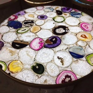 May include: A round table with a surface made of white, oval-shaped stones inlaid with colorful agate slices. The agate slices feature shades of purple, blue, green, yellow, and pink. The table has a gold-colored metal rim.