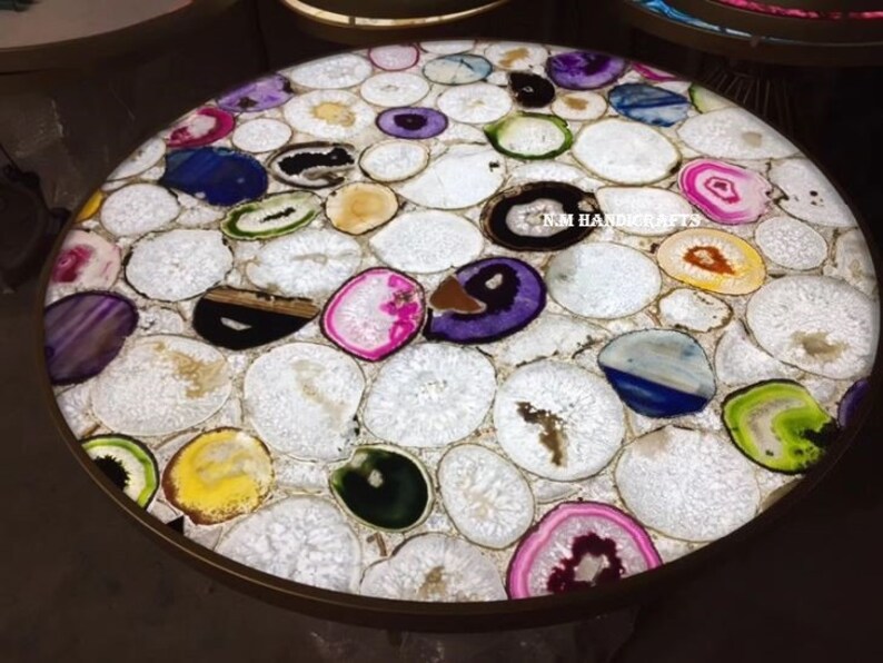 May include: A round table with a surface made of white, oval-shaped stones inlaid with colorful agate slices. The agate slices feature shades of purple, blue, green, yellow, and pink. The table has a gold-colored metal rim.