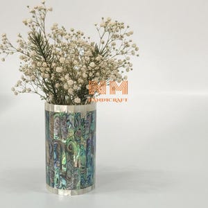 Peut inclure: Un vase cylindrique avec un motif en mosaïque de nacre dans des tons de vert, de bleu et de reflets irisés. Le vase est rempli de délicates fleurs blanches. Les bords supérieur et inférieur sont argentés. Le texte "HANDICRAFT" est visible.