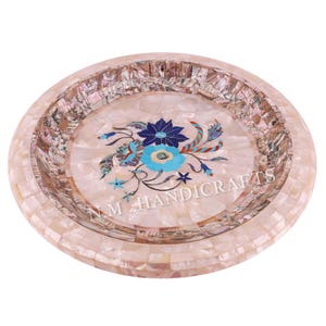 May include: A round, white, decorative plate with a floral design made of inlaid mother of pearl and semi-precious stones. The plate has a raised rim and a floral design in the centre. The floral design features blue, turquoise, and red flowers with green leaves. The plate is made by N.M. Handicrafts.