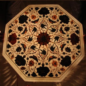 May include: Octagonal table top featuring an intricate floral design inlaid with various coloured stones. The design incorporates birds, flowers, and leaves in shades of black, white, brown, red, and green.