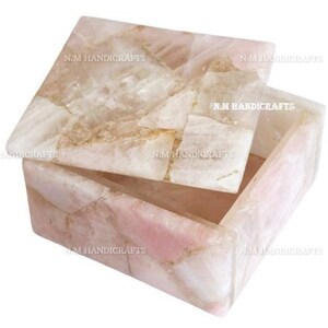May include: A square, open rose quartz box with a lid slightly ajar. The box is made of light pink and white stone pieces, with visible veining and a natural, textured appearance. The lid is also made of the same stone. The text "N.M HANDICRAFTS" is visible on the box.