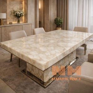May include: A rectangular dining table featuring a polished cream-colored stone surface with a natural marbled pattern. The table is supported by a thick, matching stone pedestal base with a metallic trim at the bottom. Text reads NM HANDICRAFT.