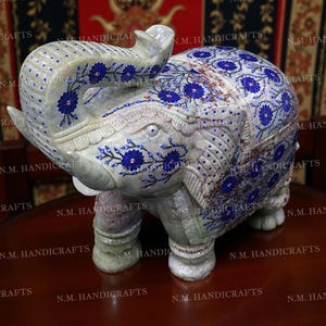 May include: A detailed, hand-carved elephant sculpture. The light-coloured stone is adorned with intricate blue floral patterns. The elephant's trunk is raised, and the sculpture has a textured surface. The base is a dark brown colour.