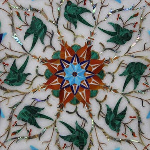 May include: A white marble table top with a decorative inlay of green, blue, red, and orange stones. The inlay features a central flower design with eight petals and a star in the centre. The design is surrounded by a repeating pattern of birds and branches.