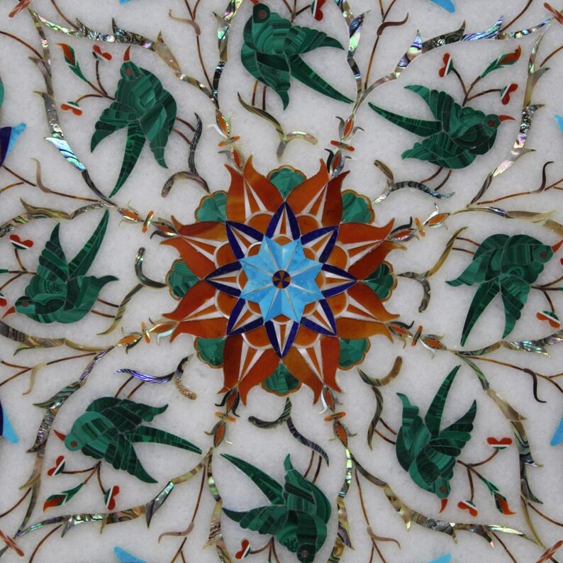 May include: A white marble table top with a decorative inlay of green, blue, red, and orange stones. The inlay features a central flower design with eight petals and a star in the centre. The design is surrounded by a repeating pattern of birds and branches.