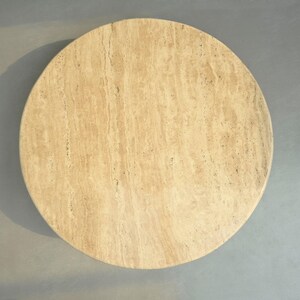May include: A round, light beige travertine table top. The surface has a natural, veined pattern.