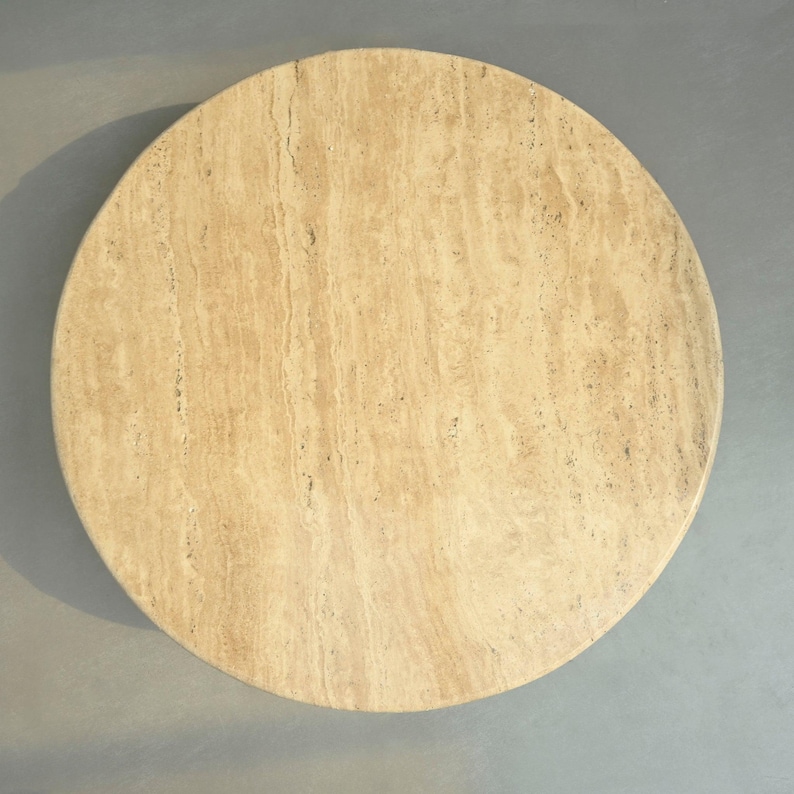 May include: A round, light beige travertine table top. The surface has a natural, veined pattern.