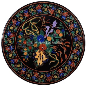 May include: A round black marble table top with a floral design inlaid with colorful stones. The design features a bouquet of flowers with green leaves and stems. The table top is surrounded by a border of smaller floral designs.