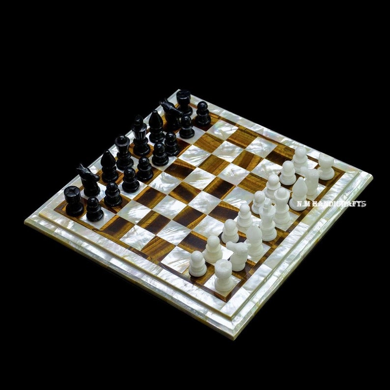 May include: A white and brown chessboard with black and white chess pieces. The board is made of mother of pearl and has a tiger eye inlay. The chess pieces are made of bone. The board is tilted at an angle, and the pieces are arranged in a starting position. The text "K.M. HANDICRAFTS" is visible in the lower right corner of the board.