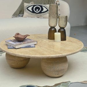 May include: A round, light brown travertine coffee table with three legs. The legs are round and have a textured surface. The table is decorated with a small brown ceramic dish, a book, and two glass candle holders with a white candle.
