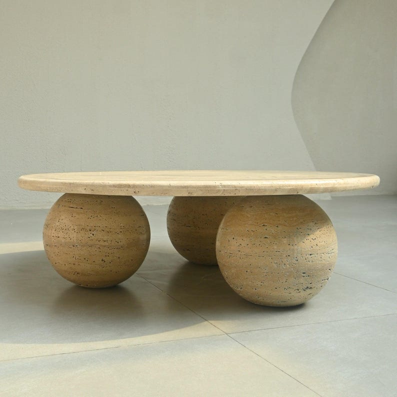 May include: A light beige oval-shaped coffee table with three round legs made of the same material. The table is made of travertine stone.