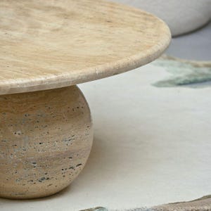 May include: A round, light brown travertine coffee table with a smooth, slightly raised edge. The table has a round, ball-shaped base.