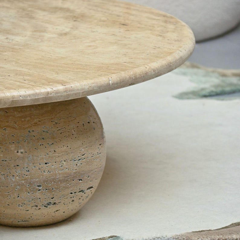 May include: A round, light brown travertine coffee table with a smooth, slightly raised edge. The table has a round, ball-shaped base.